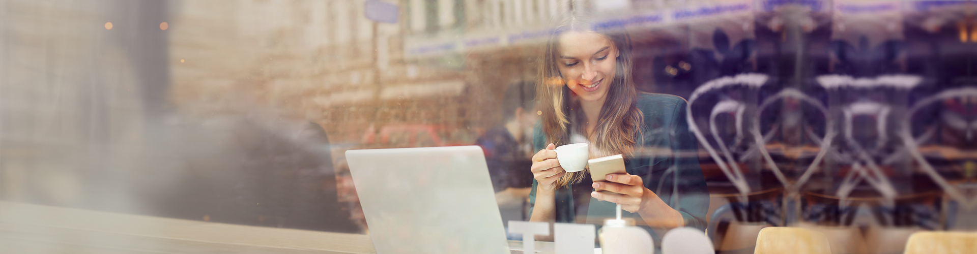 Uśmiechnięta kobieta korzysta z laptopa i smartfona w kawiarni, trzymając filiżankę